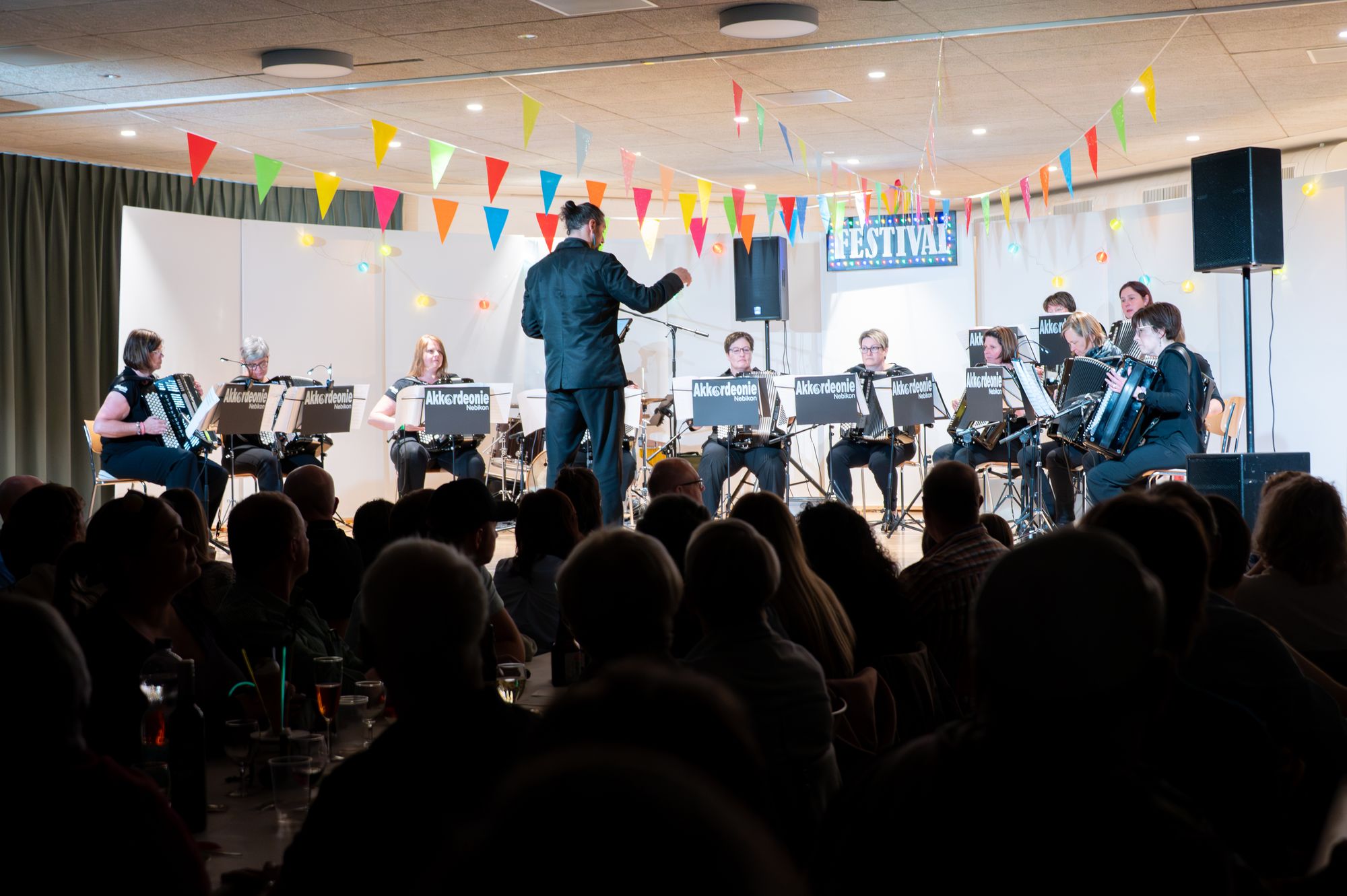 Die Akkordeonie Nebikon am Festival-Konzert: das Ensemble in Schwarz auf der Bühne, der Dirigent davor, bunte Wimpel über dem Publikum.